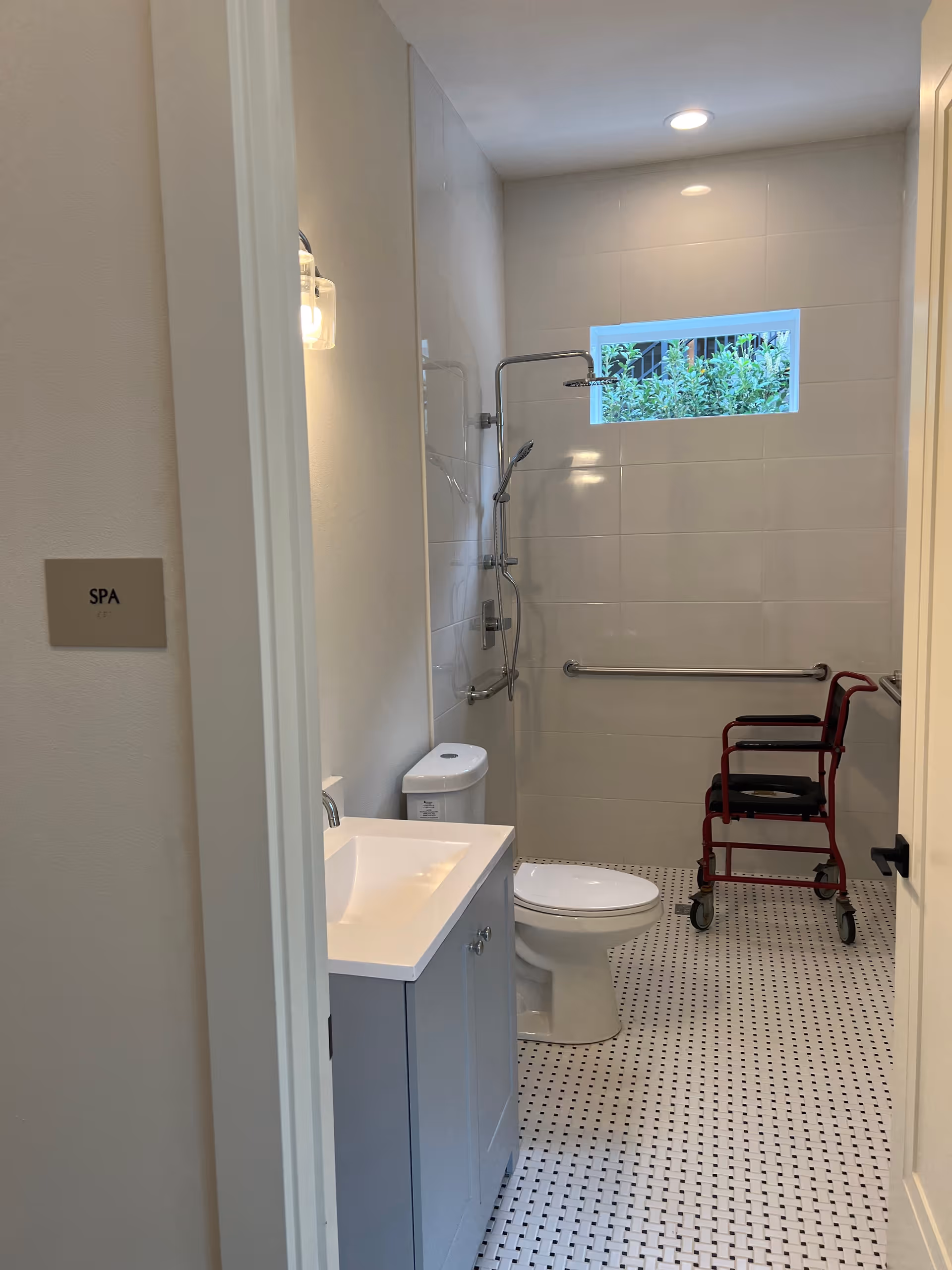 A clean, accessible bathroom with a white toilet, a gray vanity with a white sink, a shower area with grab bars, and a red shower chair. The floor has a white and black tile pattern, and there is a small window letting in natural light. A sign on the wall outside the bathroom reads 'SPA'.