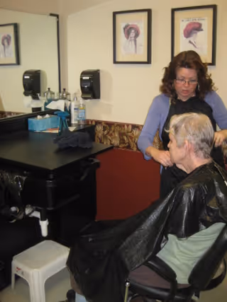 A woman is cutting the hair of an elderly person seated in a chair inside a room with a black sink and counter. The elderly person is wearing a black hairdressing cape. There are two framed pictures on the wall and a tissue box and hair care products on the counter.