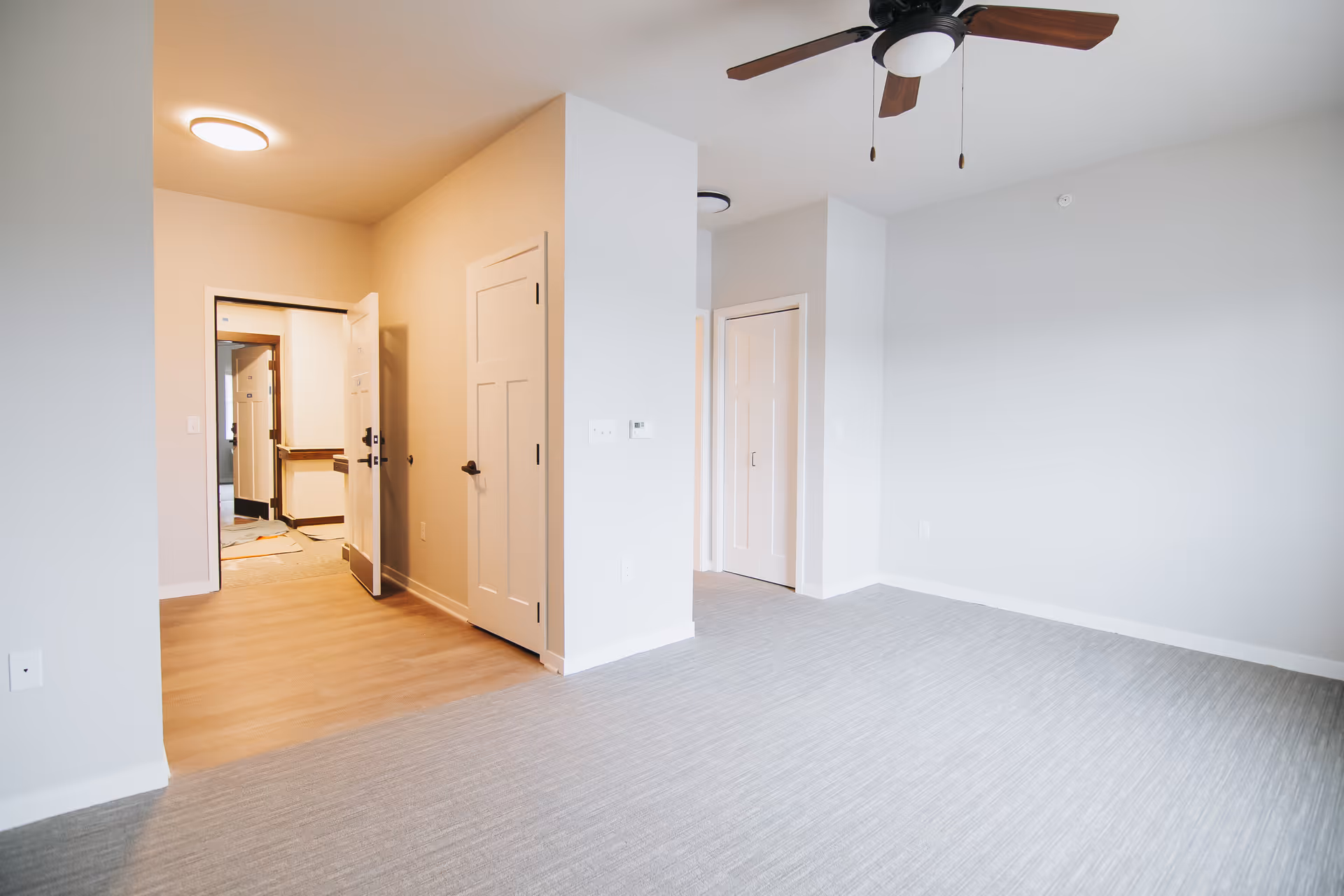 Empty light-filled room with a ceiling fan, closet doors, and an open doorway leading to a hallway.