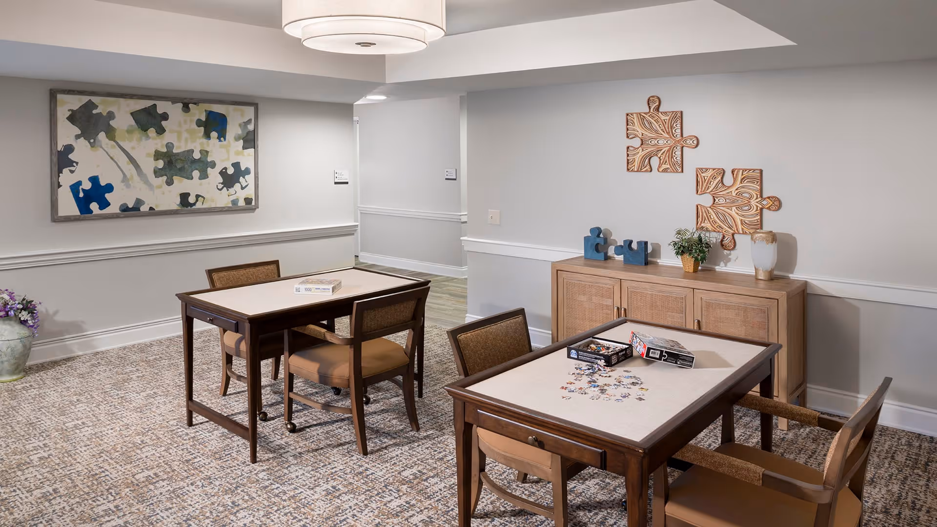 A well-lit interior room with two tables and chairs arranged for activities. One table has a partially completed jigsaw puzzle with puzzle pieces scattered on it, and the other table has a closed puzzle box. The walls are decorated with puzzle-themed artwork, including a large framed picture of puzzle pieces and two wooden puzzle piece decorations mounted above a wooden cabinet with a small plant and vase on top. The room has a patterned carpet and a modern ceiling light fixture.