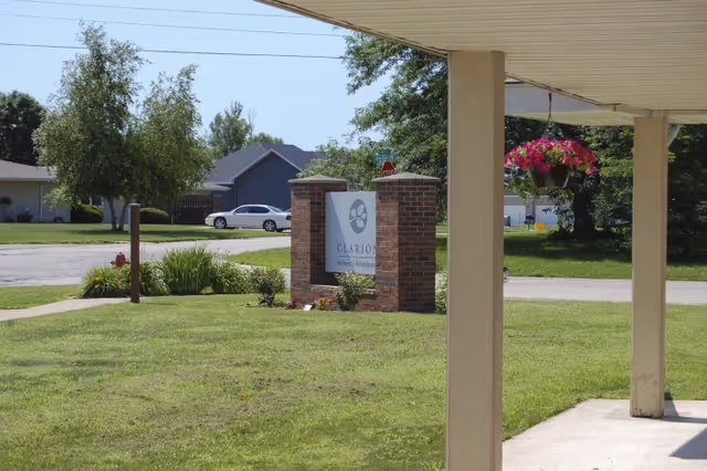 View of the outdoor area at Clarion Wellness & Rehabilitation showing a brick sign with the facility's name, a grassy lawn, a hanging flower basket, and residential houses in the background under a clear sky.