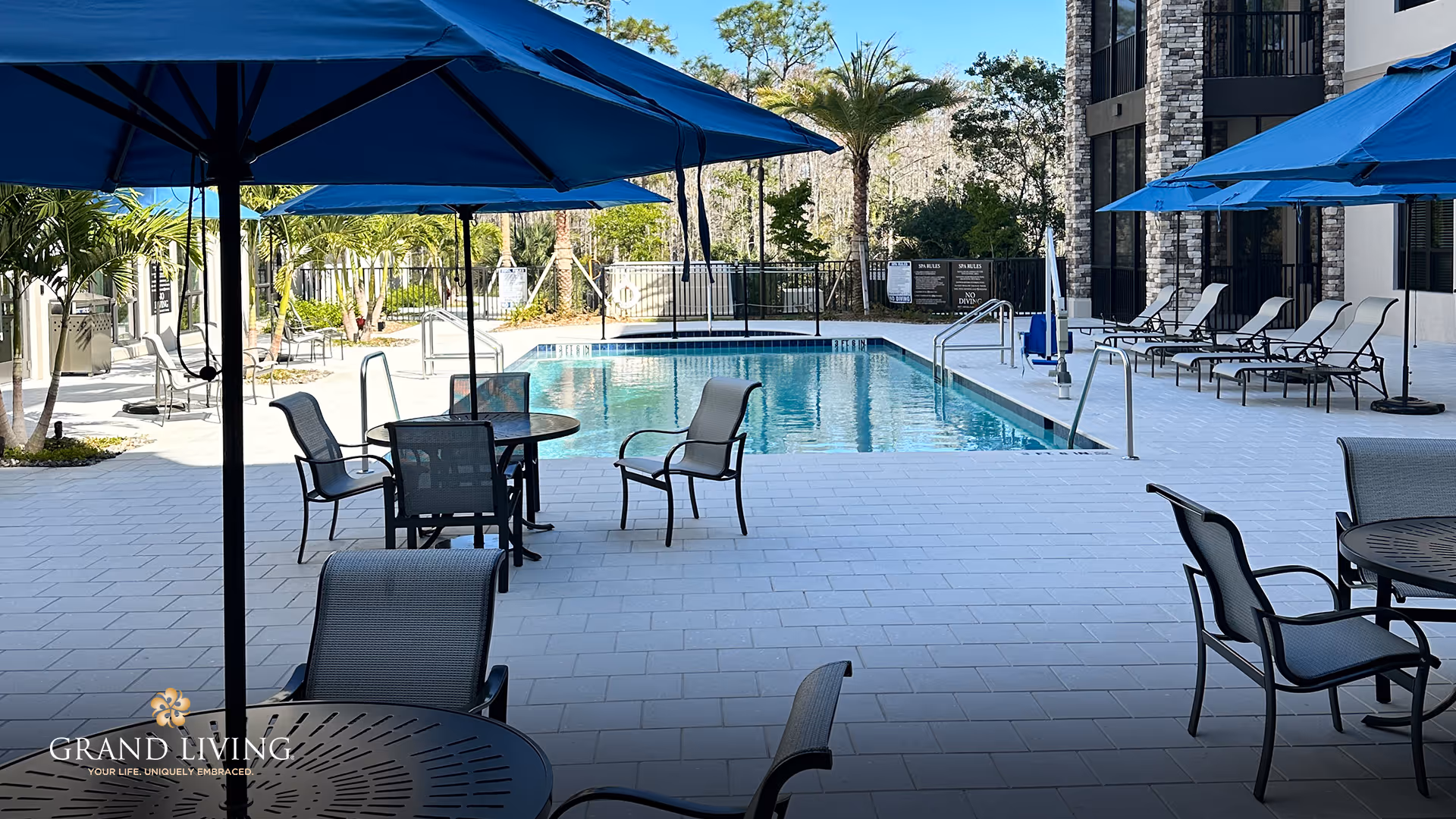 Outdoor pool area with several lounge chairs and tables with blue umbrellas surrounding the pool. Palm trees and other greenery are visible in the background, along with a multi-story building on the right side.