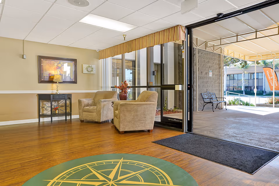A bright and welcoming seating area inside Anaheim Villa with two beige armchairs, a small black side table with a lamp, and framed artwork on the wall. The area has wooden flooring with a green and yellow compass design near the entrance. The entrance door is open, leading to an outdoor covered area with a bench and visible greenery.