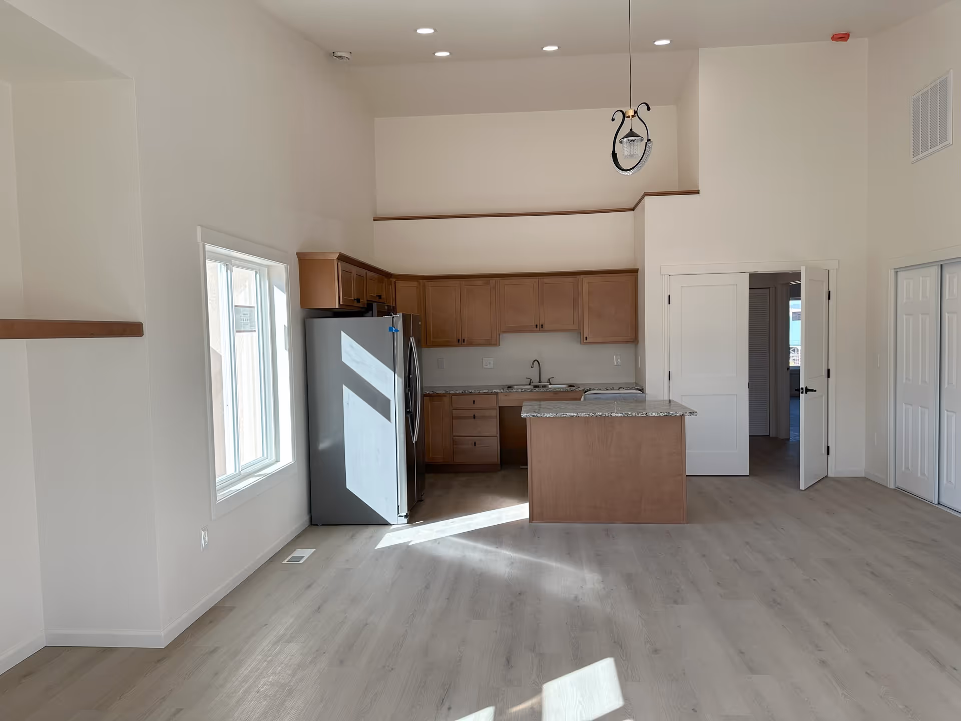 Open-concept interior showing a kitchen with a central island, stainless steel refrigerator, wood cabinets, and a bright living area.