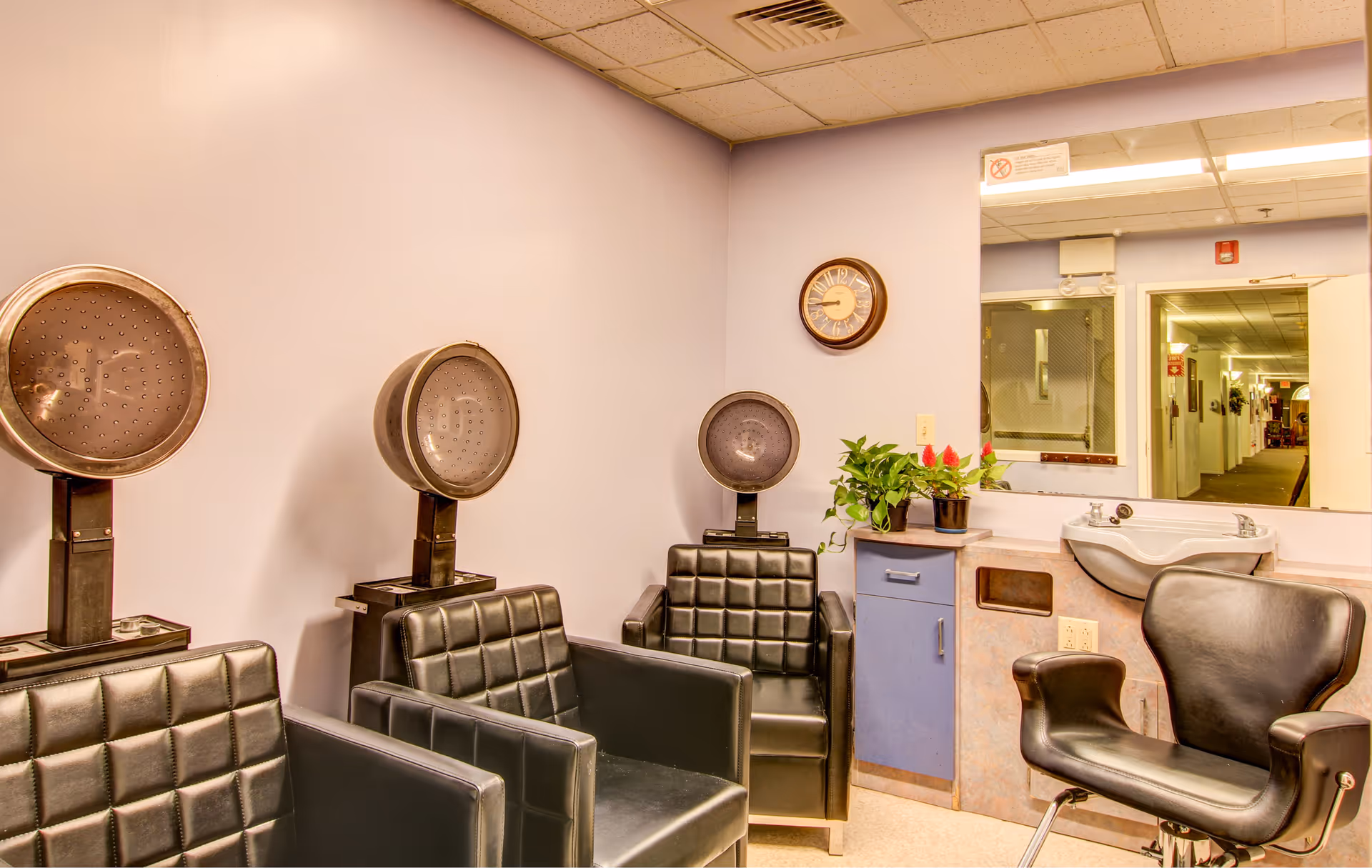 Interior of a hair salon area with three black leather chairs under hair dryers, a black salon chair in front of a sink, a wall clock, a large mirror, and a small cabinet with plants on top.