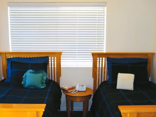 A bedroom with two twin beds separated by a small round wooden nightstand. Each bed has a wooden headboard and is made with dark blue and black checkered bedding. The left bed has a green decorative pillow, and the right bed has a white decorative pillow. On the nightstand, there is a white telephone and a book. A window with closed white blinds is above the beds.