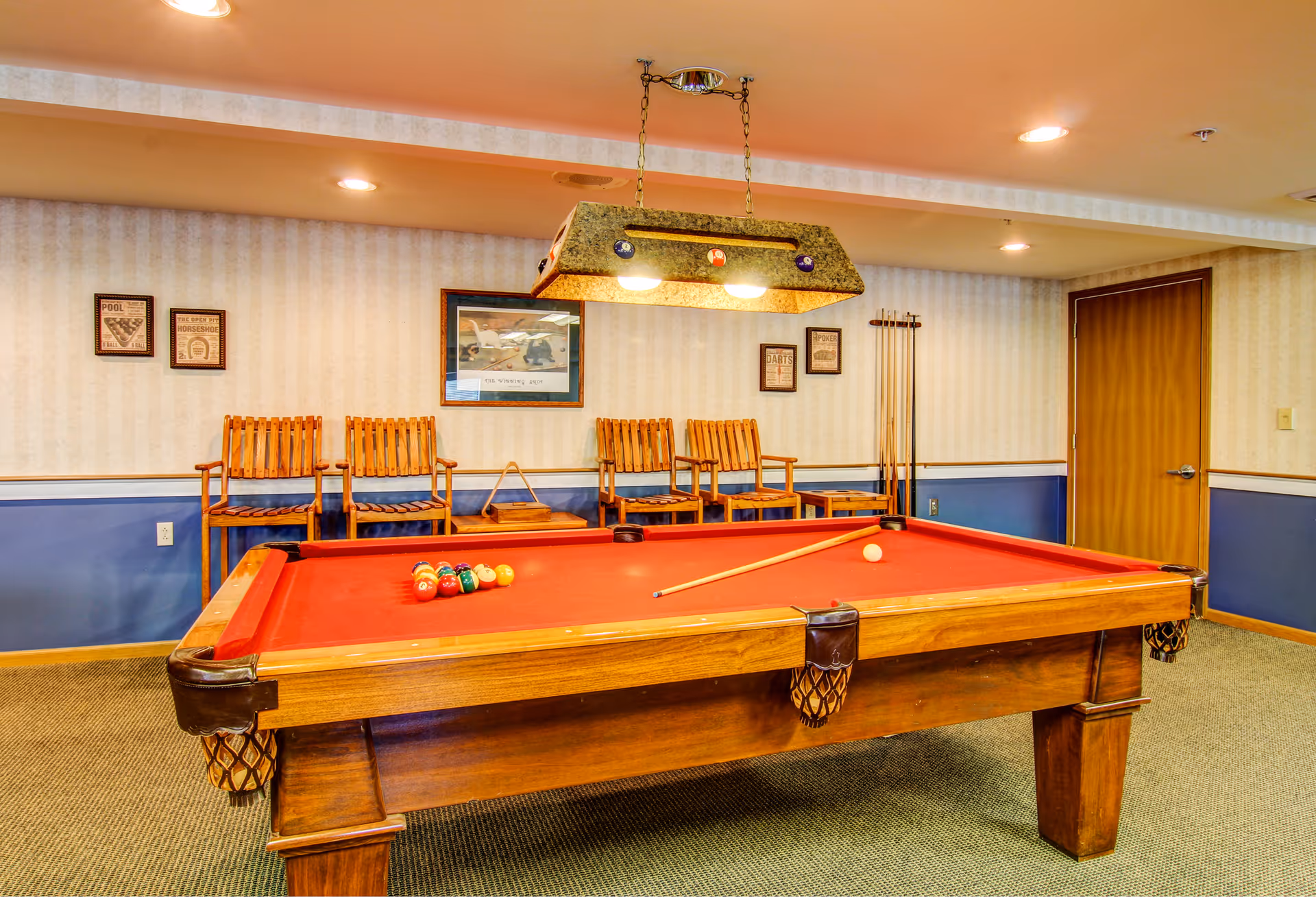 A red-felt pool table in a game room with wooden chairs along the back wall and a hanging light fixture above.