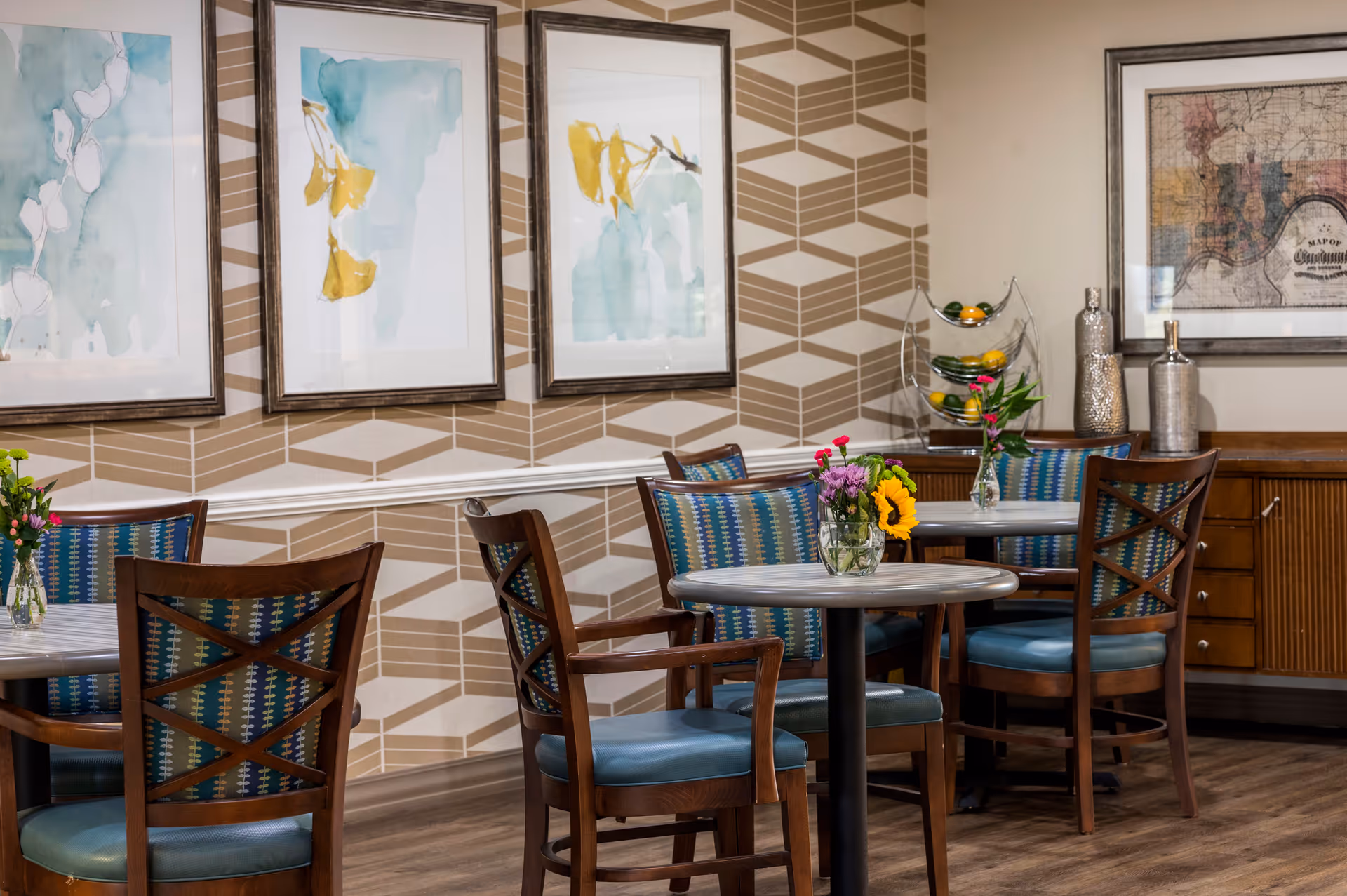 A cozy dining area with round tables and wooden chairs featuring blue patterned cushions. Each table has a small vase with colorful flowers. The walls are decorated with geometric patterned wallpaper and framed abstract artwork. A wooden sideboard with decorative vases and a fruit stand is visible in the background.