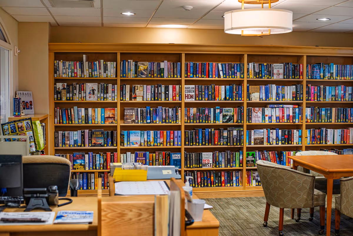 Interior common room with a large wall of bookshelves, a desk in the foreground, and a table with chairs.