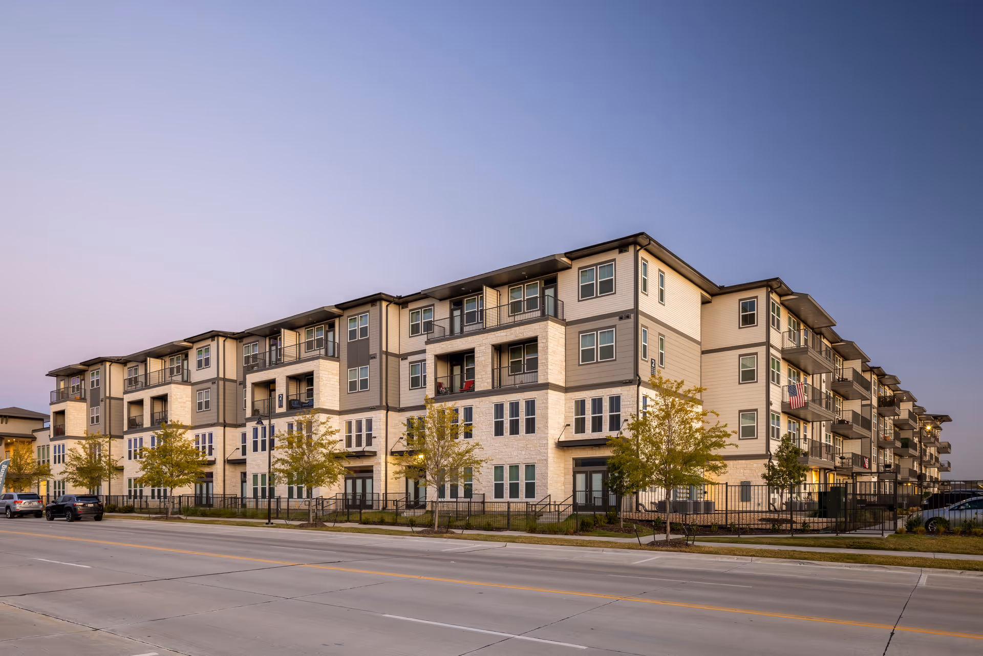 Exterior view of a modern multi-story residential building with balconies, trees along the sidewalk, and a clear evening sky.