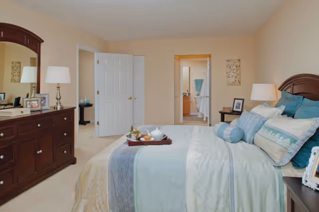 A neatly made bed with multiple pillows and a tray with a teapot and cup in a senior living community bedroom. The room features beige walls, a wooden dresser with a mirror, two bedside tables with lamps, and an open door leading to a bathroom.