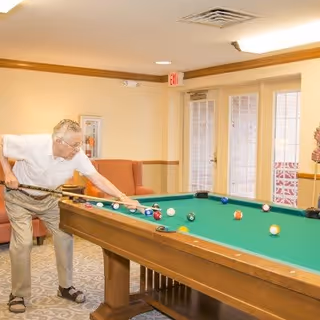 An elderly man playing pool in a well-lit room with beige walls and carpeted floor. The room has a pool table with balls arranged on it, a red couch in the background, and glass doors leading outside.
