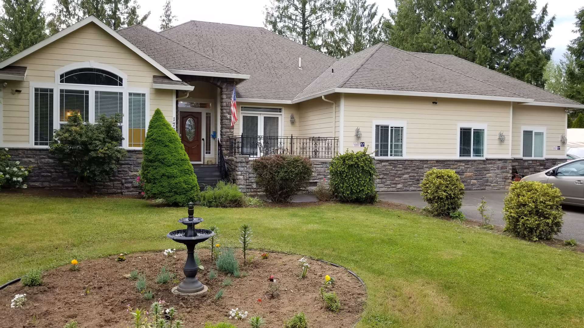 Exterior view of a single-story residential building with beige siding and stone accents. The building has multiple windows with white frames, a front door with a small porch, and an American flag hanging near the entrance. In front of the building is a well-maintained lawn with a circular flower bed containing a black tiered fountain and some small plants. There are several bushes and trees around the property, and a silver car is partially visible on the right side.