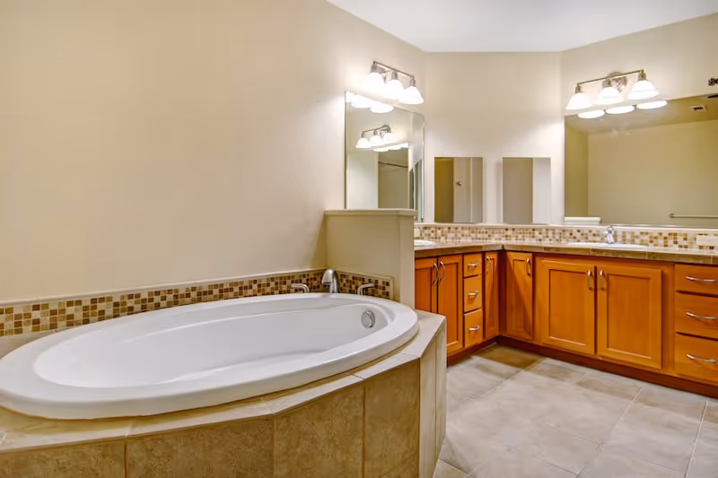A spacious bathroom featuring a large oval bathtub with tiled surround and a vanity area with wooden cabinets, a countertop, and two mirrors with light fixtures above them.