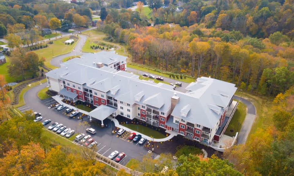 Aerial view of a large senior living facility named Keystone Place at Newbury Brook surrounded by trees with autumn foliage. The building has multiple floors with a gray roof and red and white exterior walls. There is a parking lot with several cars parked in front of the building and a driveway leading to the entrance.