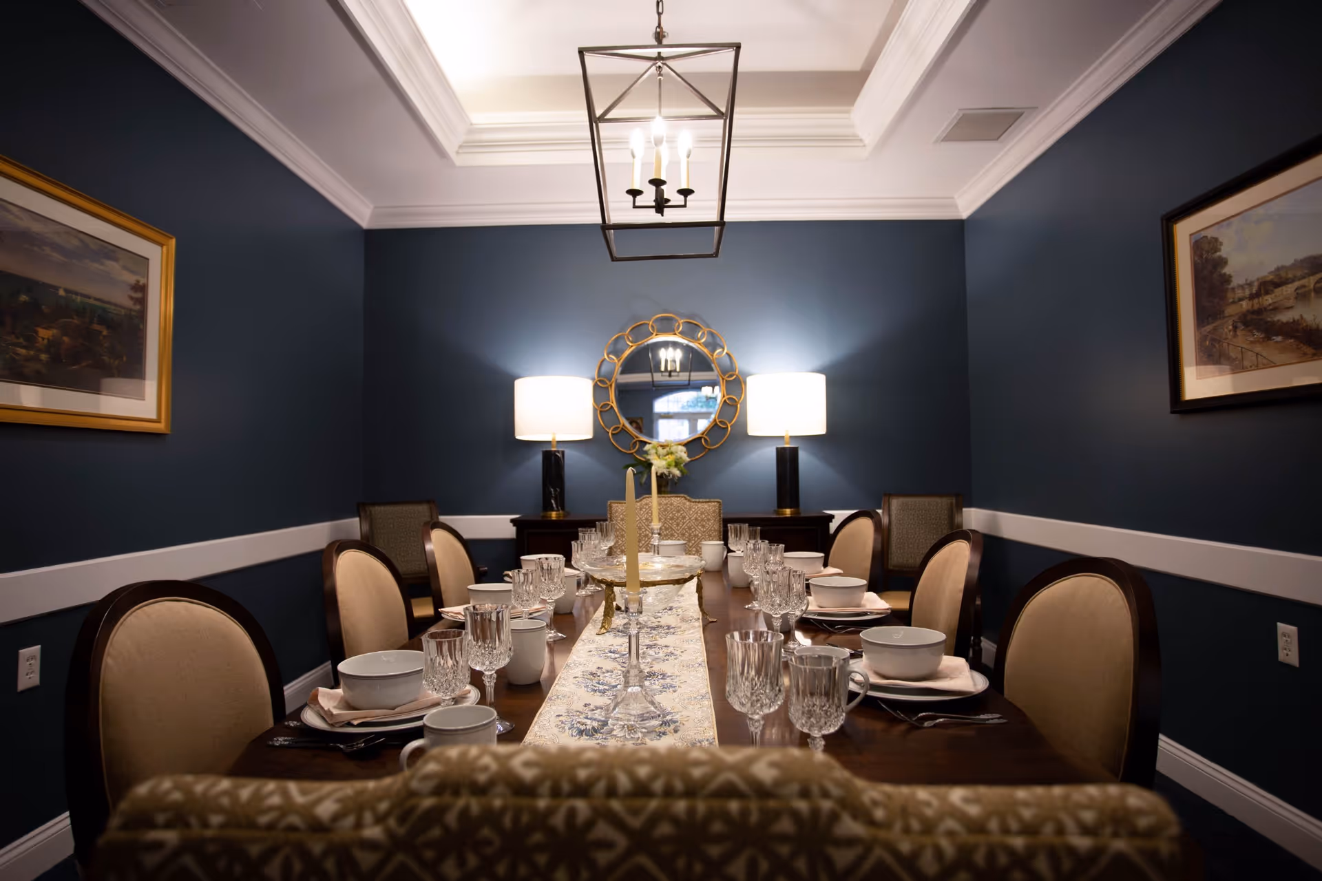 Formal dining room with a long set table, upholstered chairs, blue walls, a decorative round mirror, and table lamps.