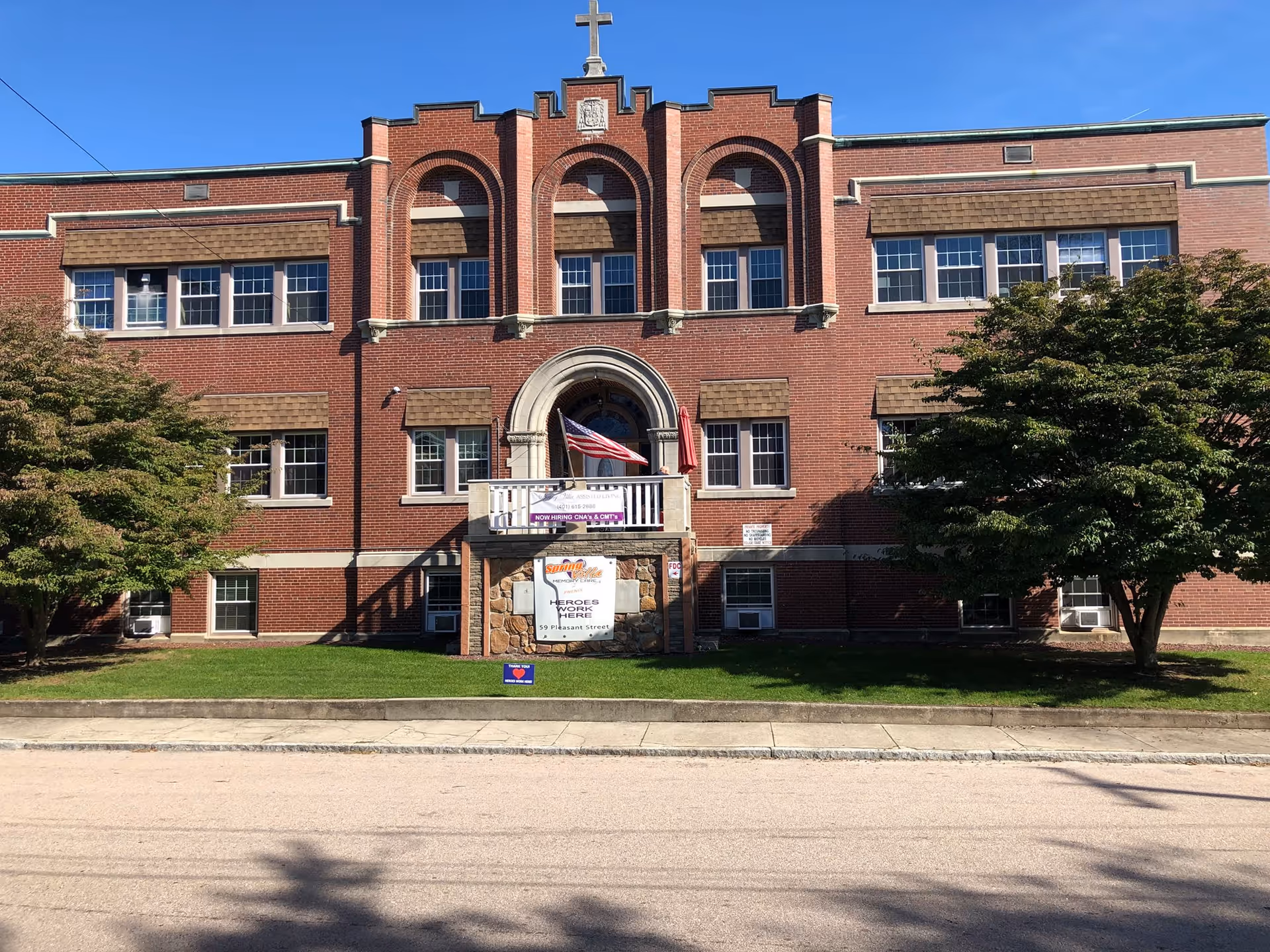 Front exterior view of a large red brick building with multiple windows, a central arched entrance with a balcony, an American flag, and signs indicating Spring Villa Memory Care and hiring information. There are two trees on either side of the entrance and a clear blue sky above.