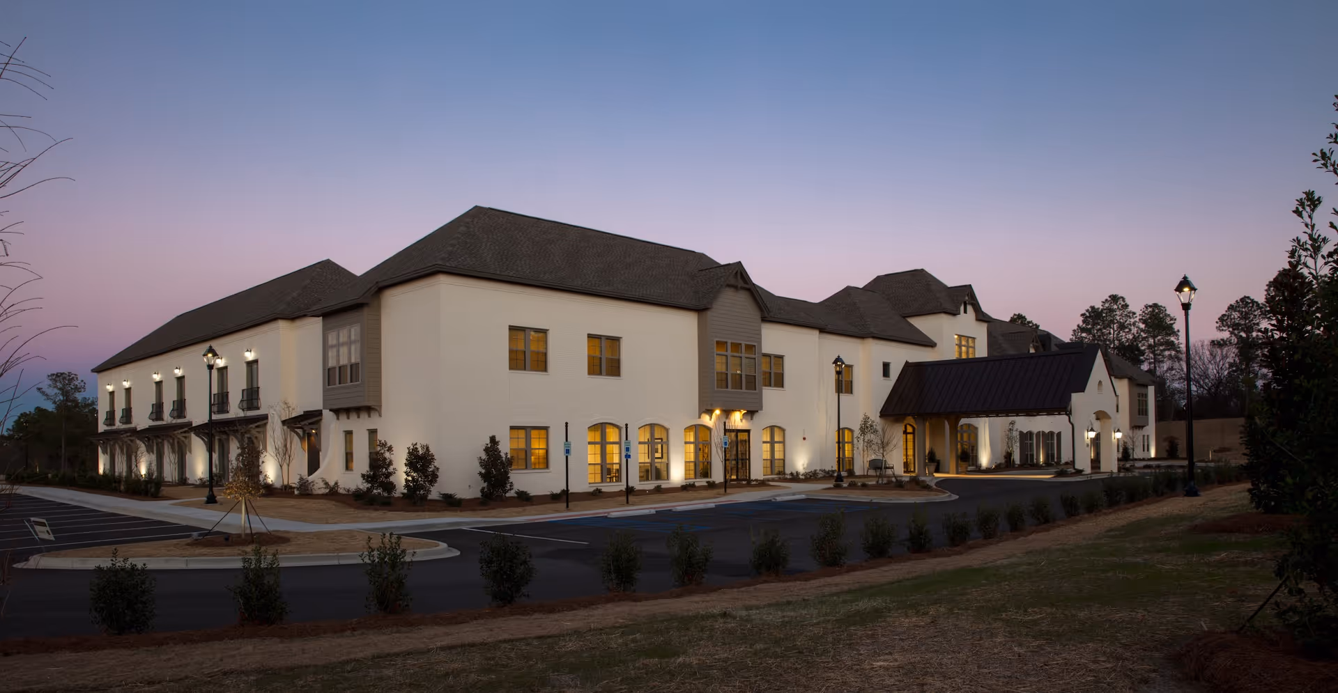 Exterior view of a large, two-story senior living facility building at dusk with lights on inside and street lamps illuminating the parking area and entrance.