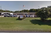 Exterior view of Autumn Care of Suffolk facility with a single-story building, a parking lot filled with cars, a flagpole with an American flag, and a green lawn with a sign displaying the facility's name.