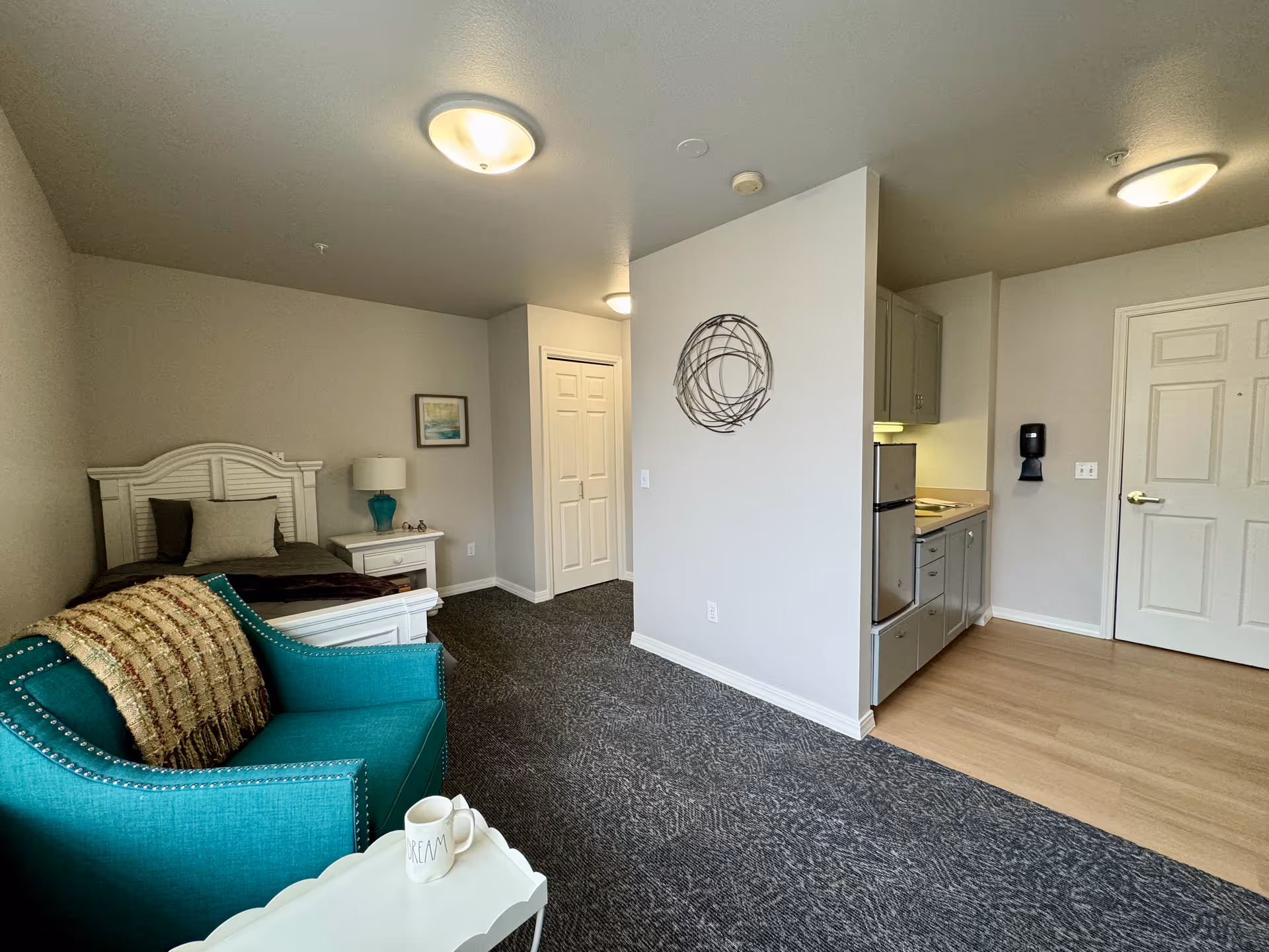 Interior view of a senior living facility room featuring a single bed with pillows and a blanket, a nightstand with a lamp and decorative items, a teal armchair with a throw blanket, a small white table with a coffee mug, a kitchenette with a mini fridge and cabinets, and a closed door. The room has carpeted and wooden flooring and neutral-colored walls with a circular wall decoration.