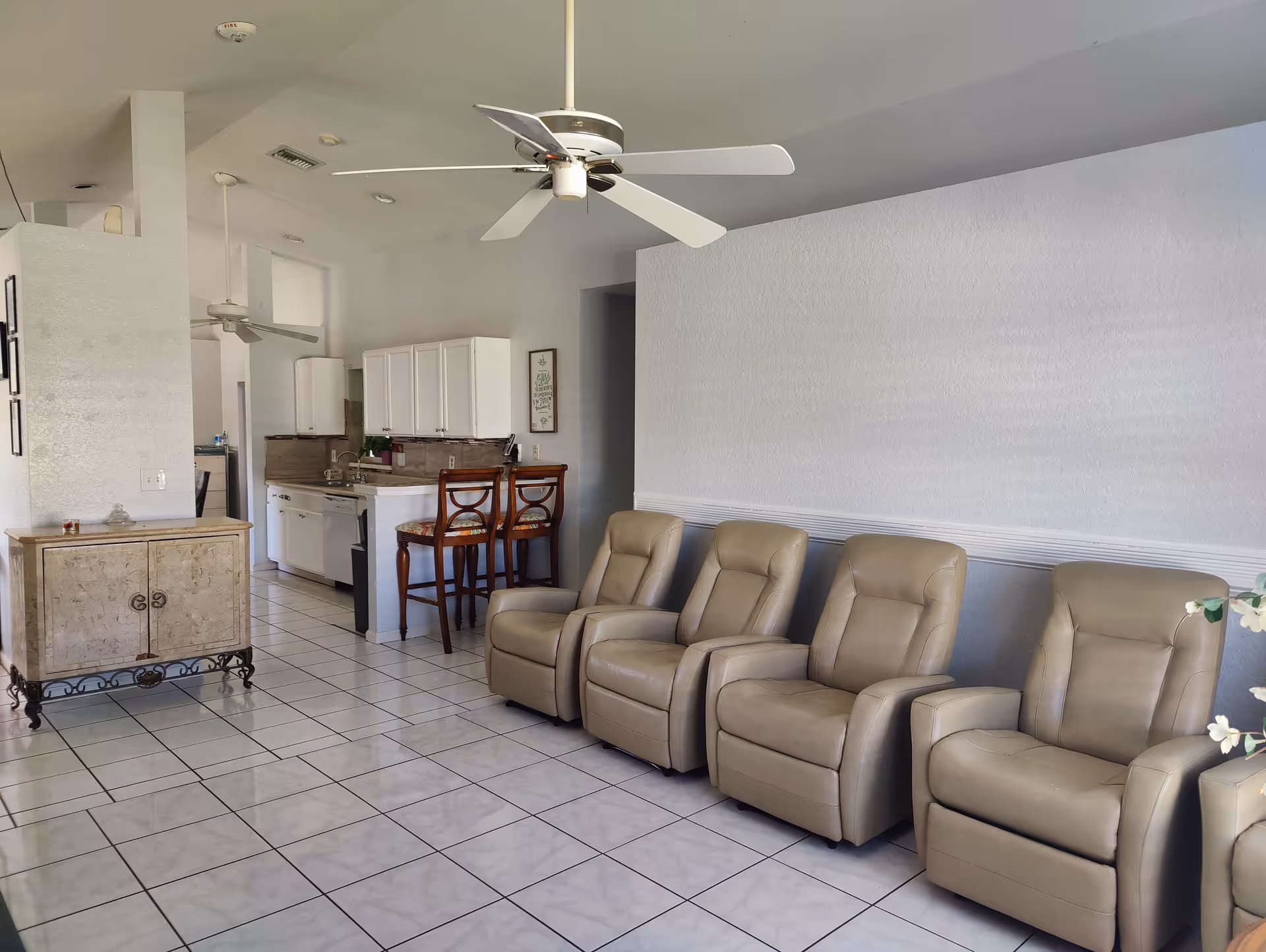 Bright common room with a row of beige recliner chairs, tile floor, ceiling fans, and a view into a white kitchen with bar stools.