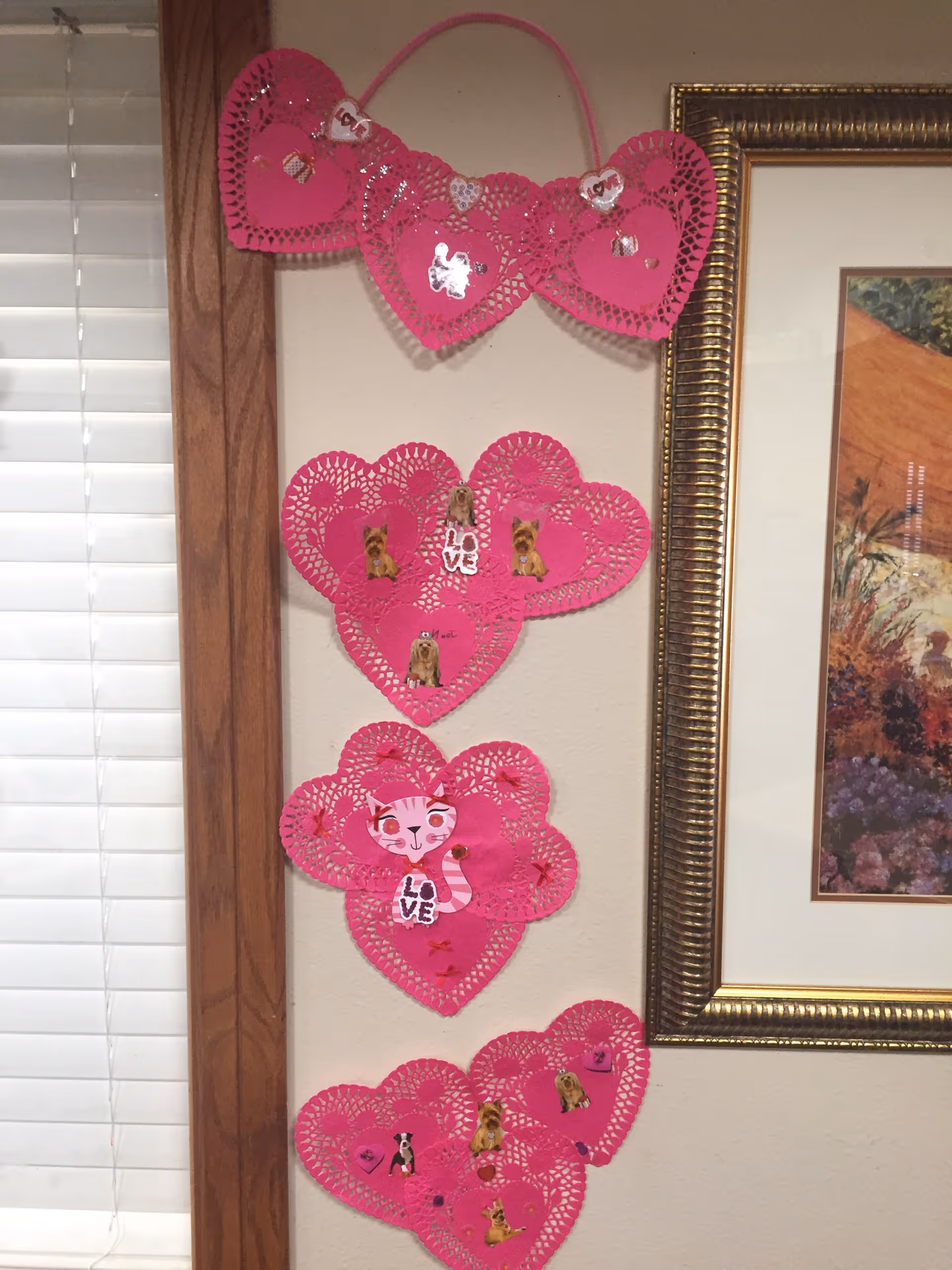 Vertical row of pink heart-shaped paper doilies with stickers hanging on an interior wall next to a framed picture and window blinds.