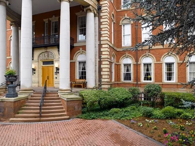 Entrance of a large brick building with tall white columns, a set of stairs leading to double wooden doors, surrounded by green bushes and a small garden with flowers.