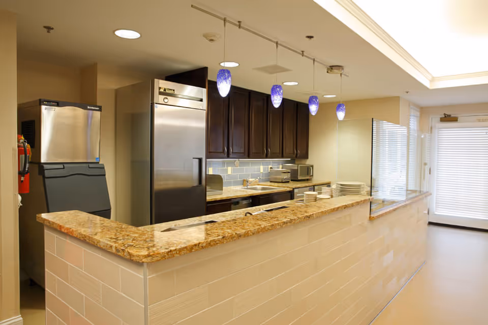 Bright serving counter and kitchenette with a granite countertop, stainless appliances, hanging pendant lights, and stacked plates.