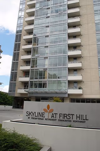 Exterior view of a multi-story building with balconies and large glass windows. In front of the building is a sign that reads 'Skyline at First Hill by Presbyterian Retirement Communities Northwest'.