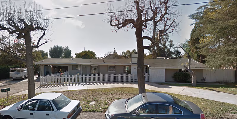 Single-story residential building with a white picket fence in front, two leafless trees on the lawn, and two parked cars on the street. The building has a light-colored exterior and a sloped roof, with some greenery around the sides.