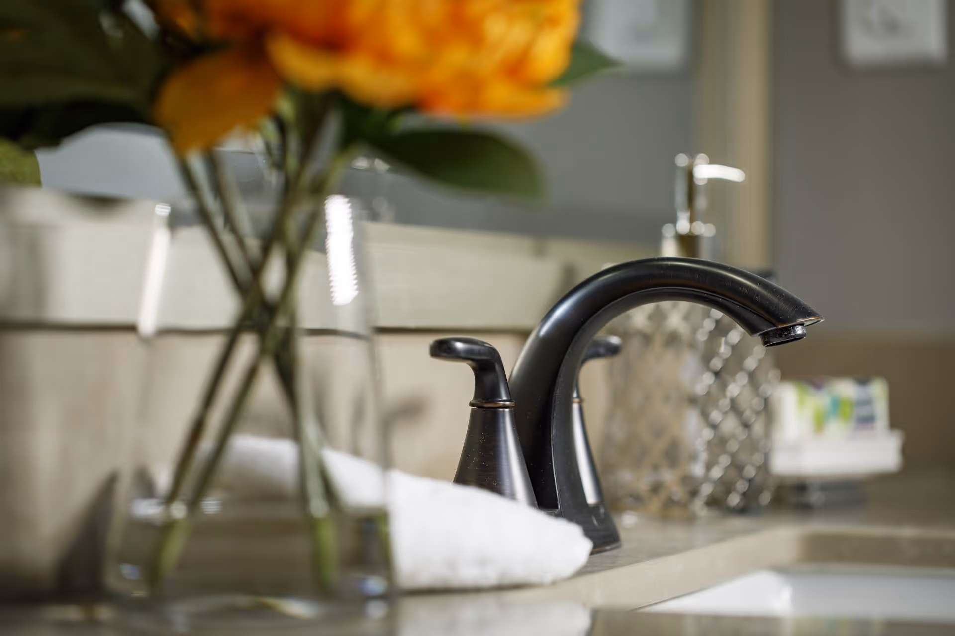 Close-up view of a bathroom sink faucet with a dark bronze finish, a white rolled towel, a glass vase with orange flowers, a soap dispenser, and a decorative container on the countertop.