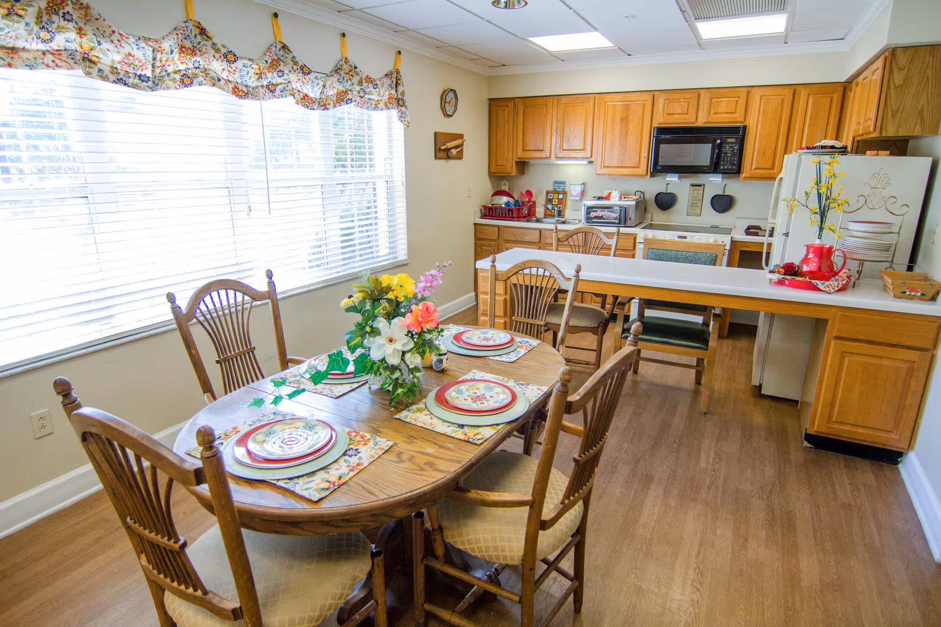A bright kitchen and dining area with wooden cabinets and a wooden dining table set for four with floral placemats and a centerpiece of colorful flowers. The kitchen has a white countertop, a microwave, a toaster oven, and a refrigerator decorated with a red pitcher and yellow flowers. Large windows with floral valances let in natural light.