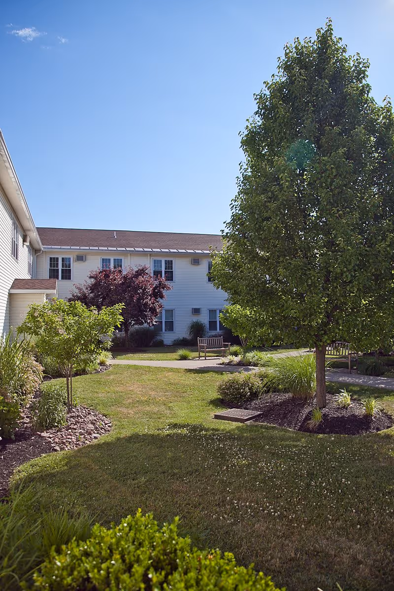 Outdoor garden area with green grass, various trees, and shrubs in front of a two-story white building under a clear blue sky. There are benches along a paved walkway in the garden.