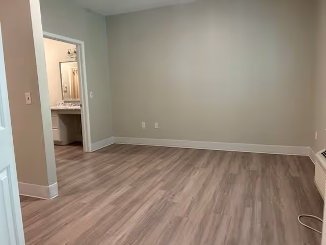 Empty room with light wood flooring and beige walls, an open doorway reveals a bathroom with a vanity and mirror.