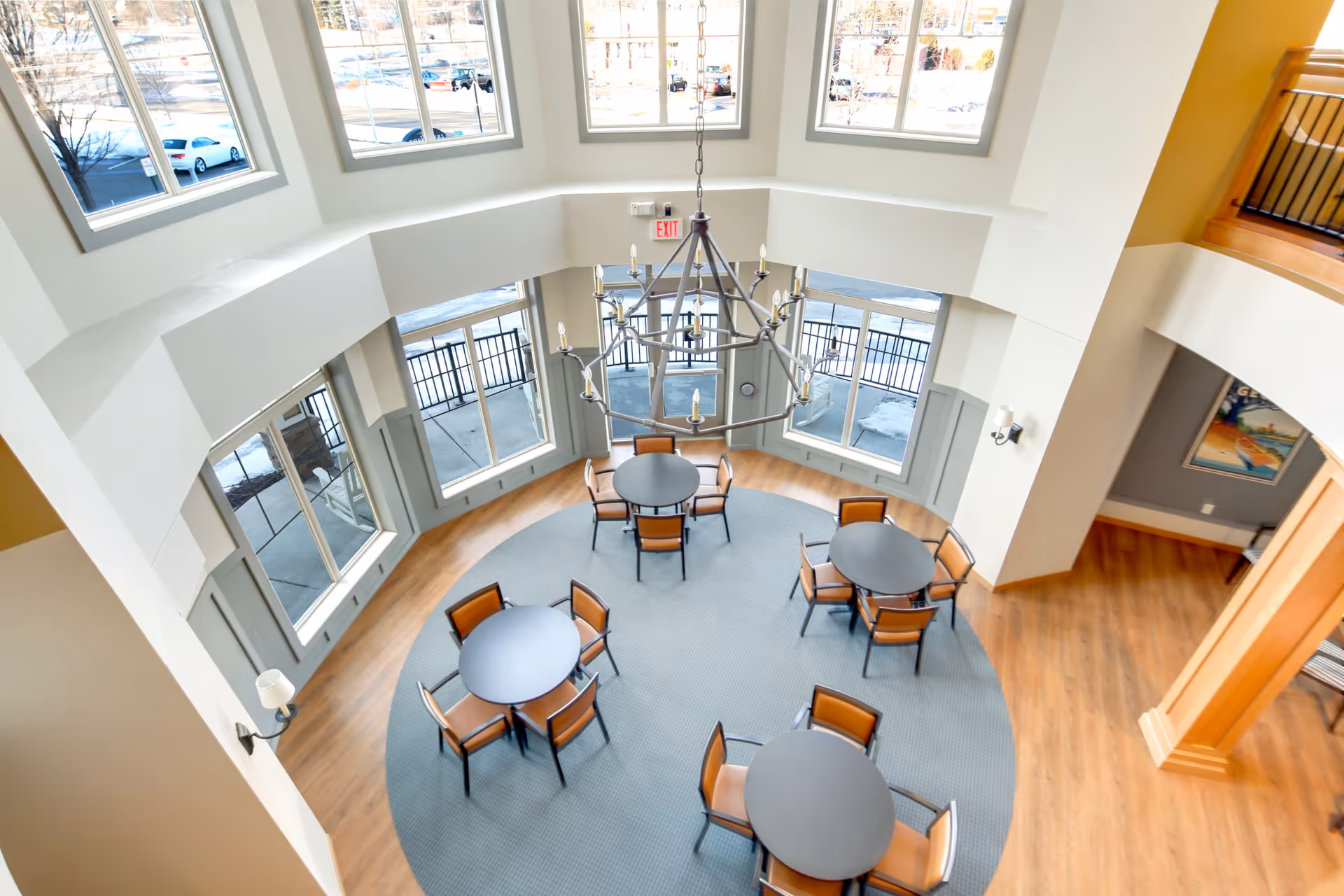 View from above of a bright, spacious common area with four round tables, each surrounded by four chairs. Large windows let in natural light and offer a view of the outside parking area. A modern chandelier hangs from the ceiling above the tables, and the floor is a combination of wood and carpet.