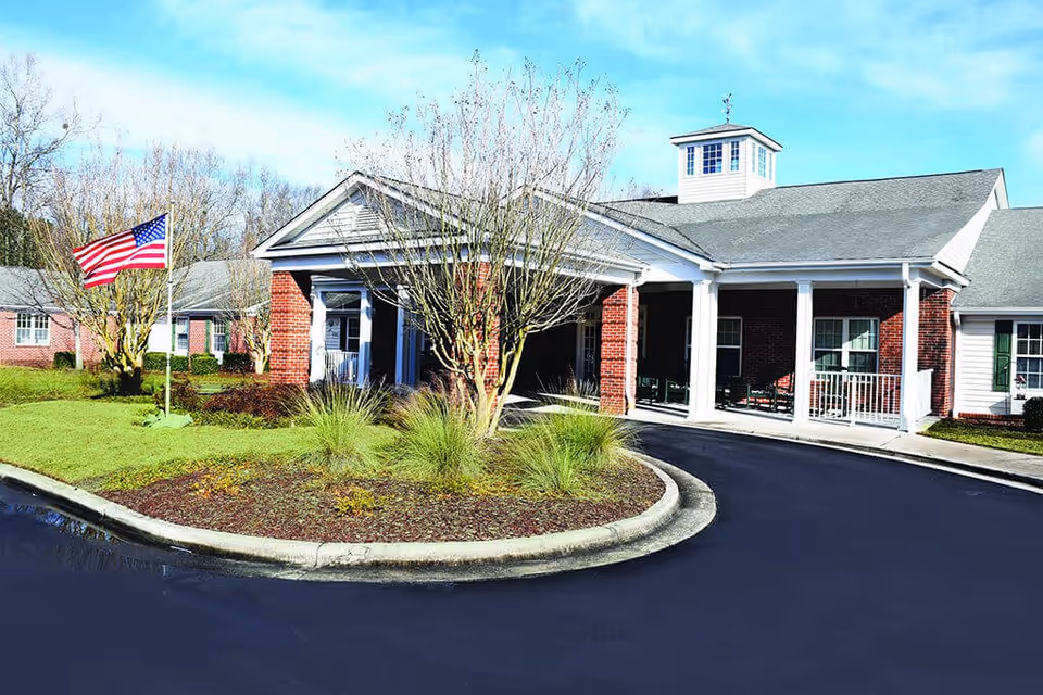Exterior view of a senior living facility building with a covered entrance, brick walls, white columns, and a small tower on the roof. There is a landscaped roundabout with grass, shrubs, and a tree in front of the entrance. An American flag is flying on a flagpole to the left of the entrance. The sky is clear and blue.