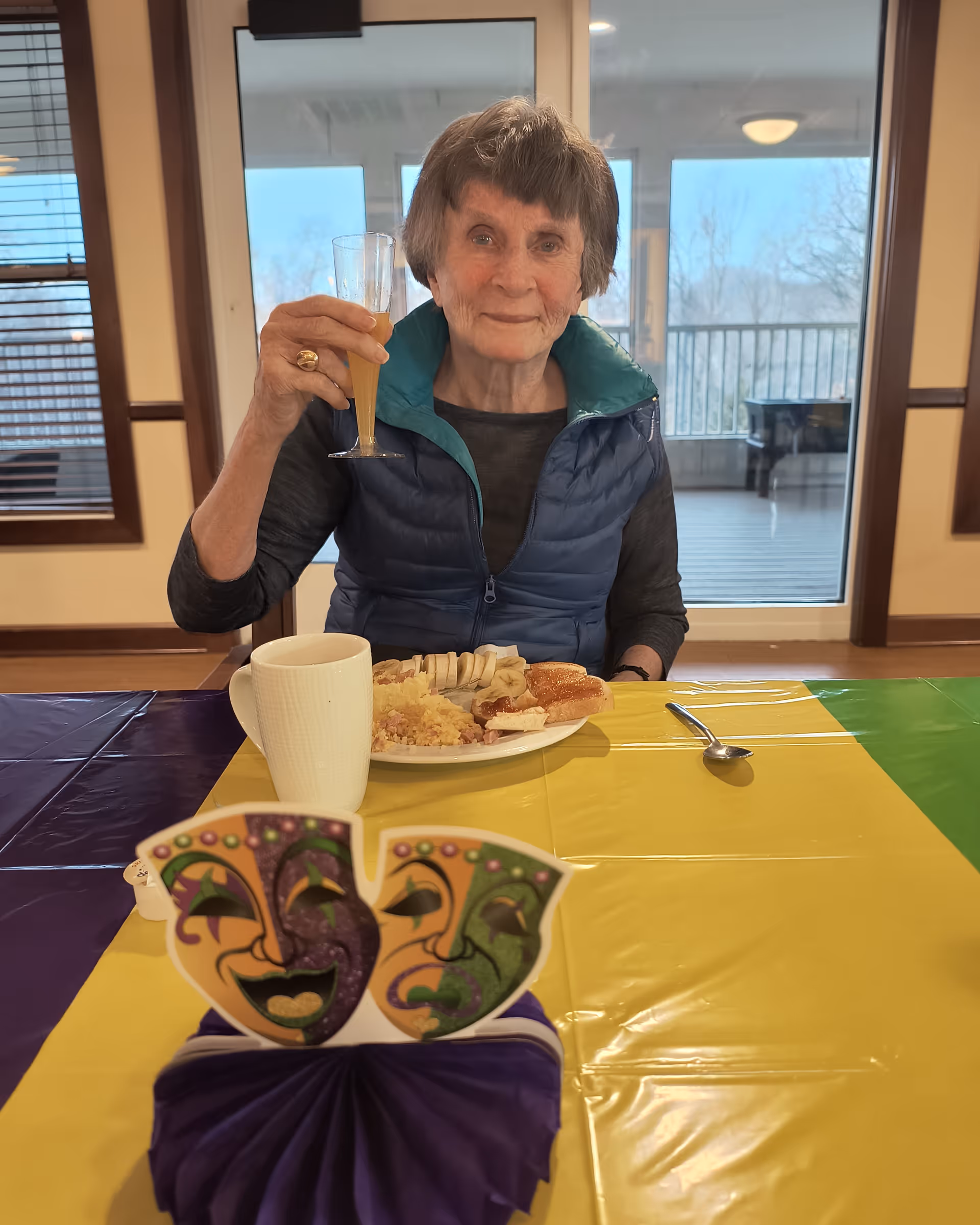 An older woman seated at a colorful, Mardi Gras-decorated dining table raising a glass with a plate of food and a mug in front of her.