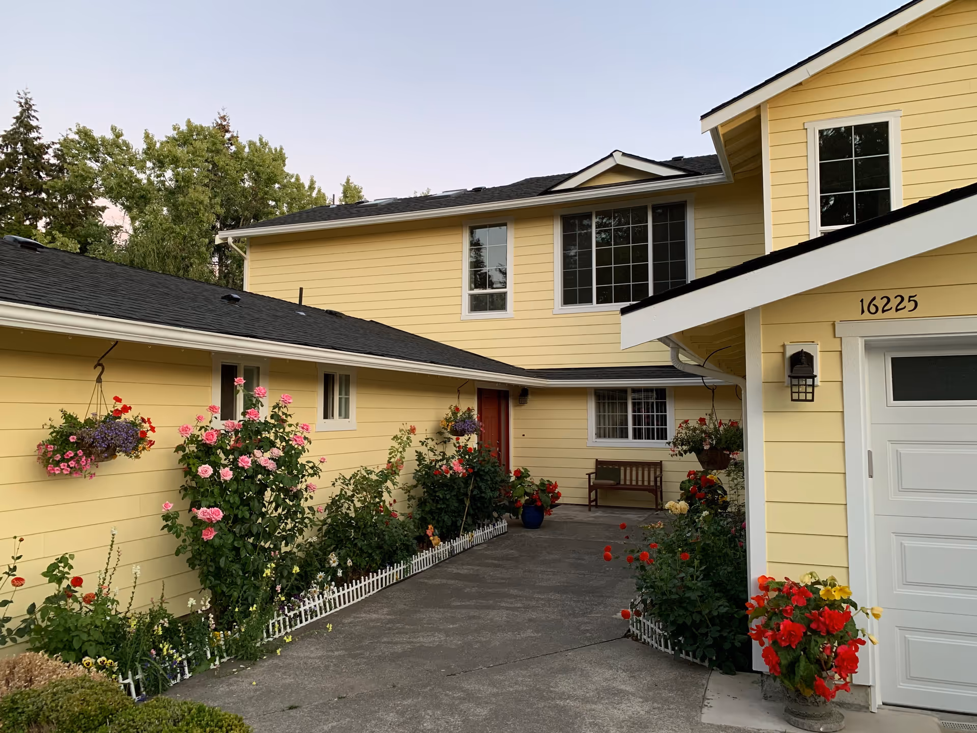 Yellow two-story house with a driveway flanked by flowering plants, a bench by the entrance, and a garage numbered 16225.