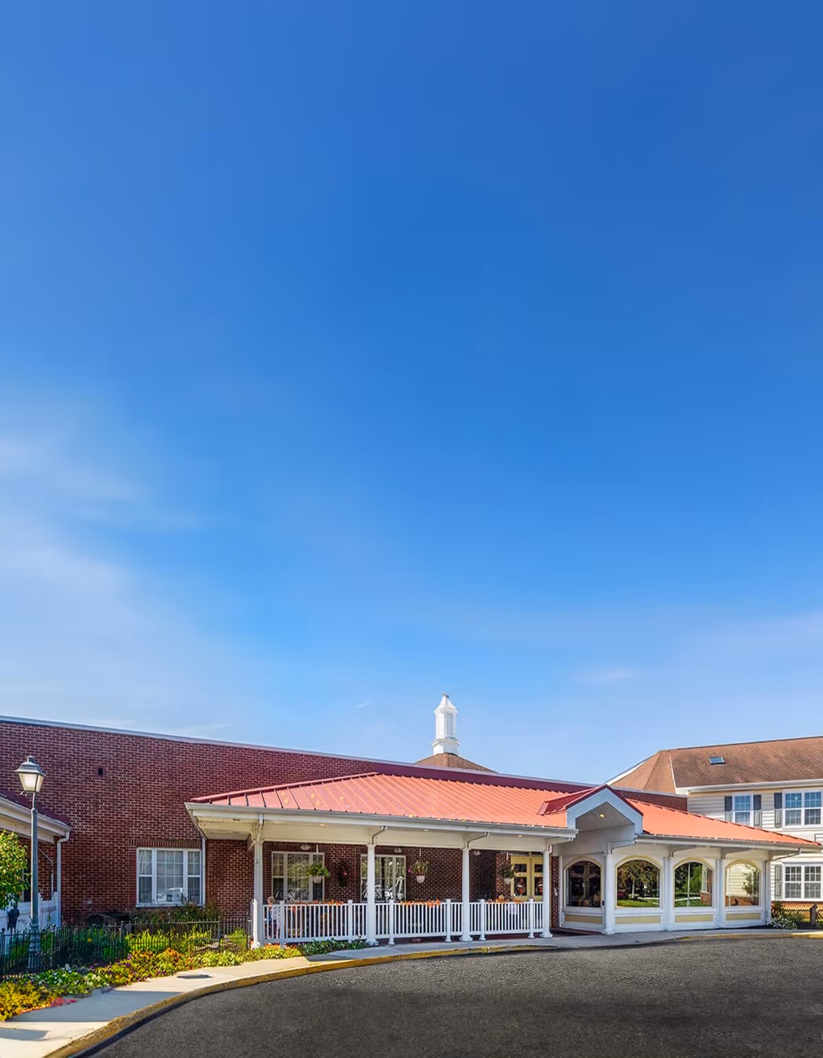 Front entrance of a red-brick senior living building with a covered porte-cochère, white railing, and a clear blue sky above.