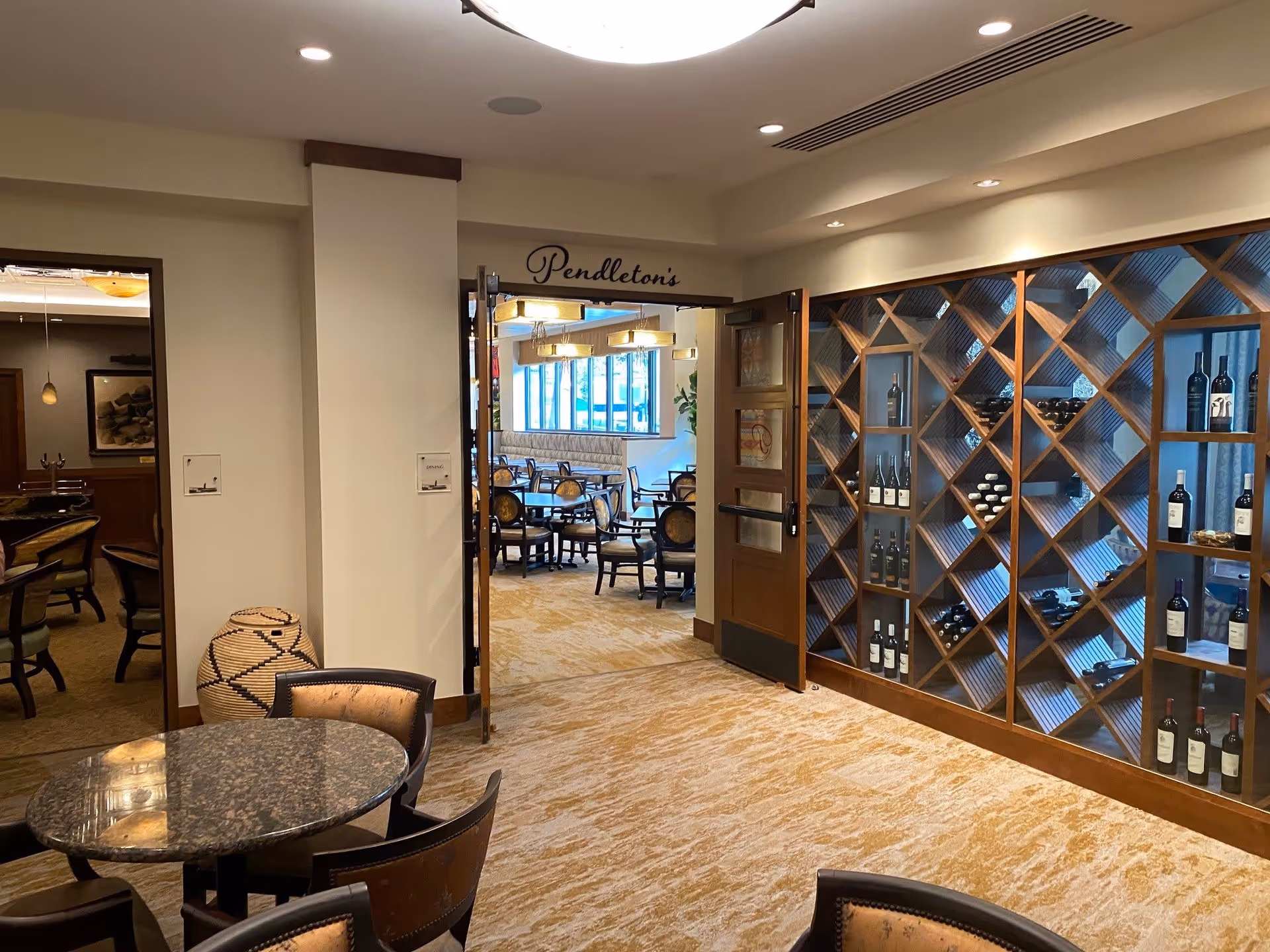 Interior view of a senior living facility showing a seating area with a round table and chairs in the foreground, a large wooden wine rack filled with bottles on the right wall, and an open doorway leading to a dining area with multiple tables and chairs. The dining area has large windows letting in natural light and a sign above the door that reads 'Pendleton's'.