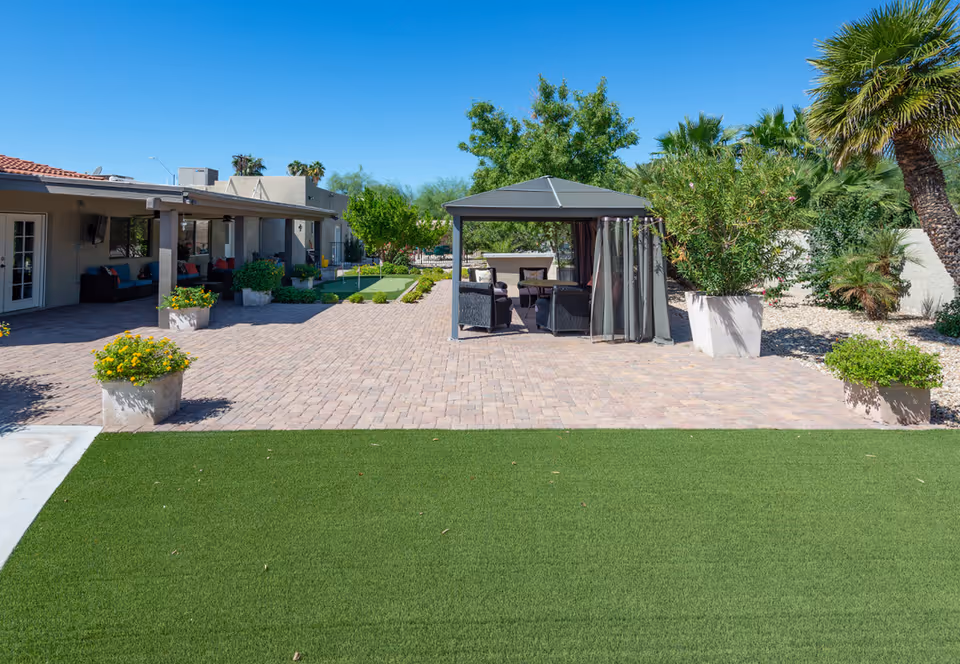 Outdoor patio area with artificial grass in the foreground, a paved brick area with a gazebo containing outdoor furniture, potted plants, and trees surrounding the space under a clear blue sky.