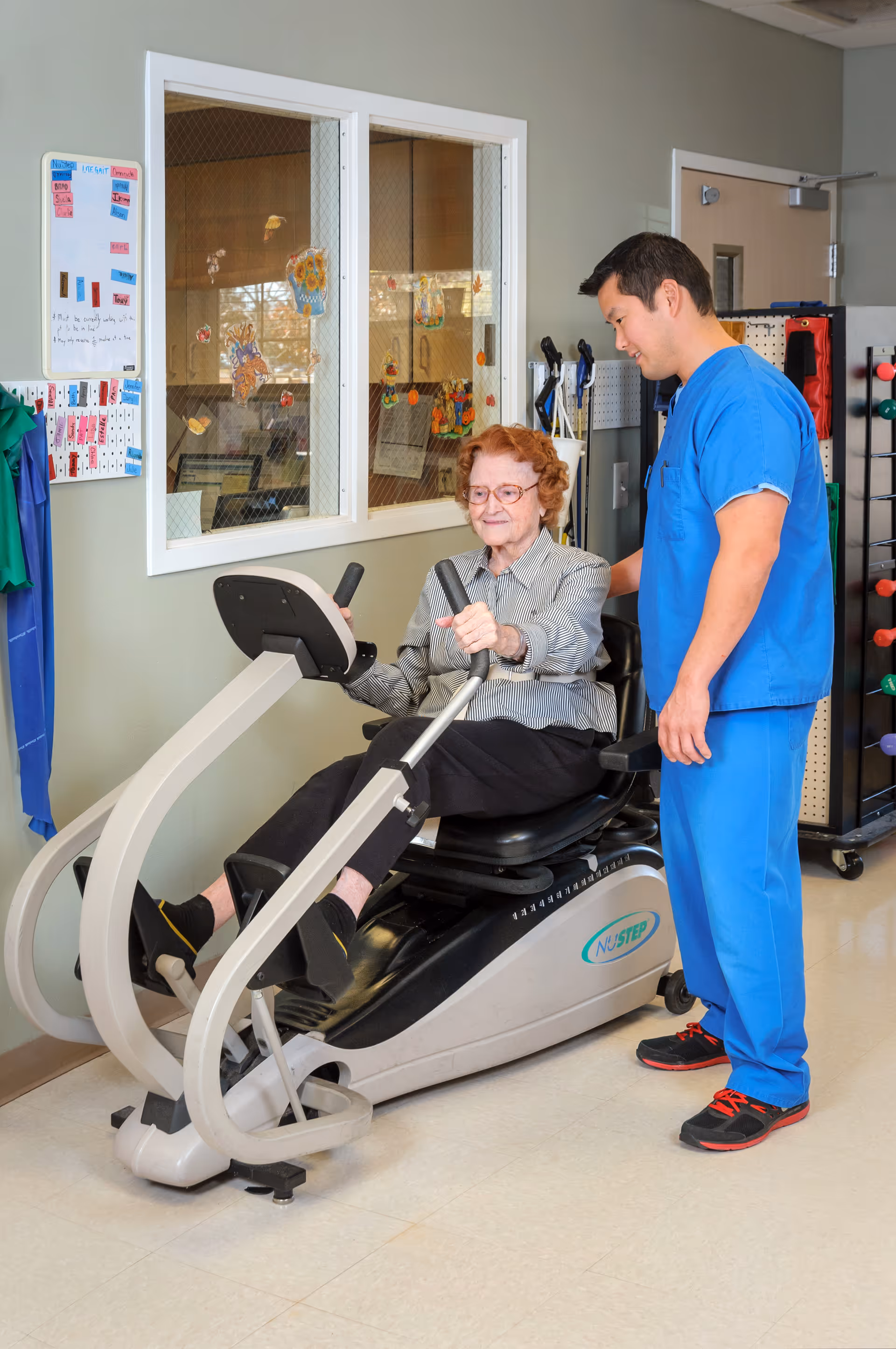 An elderly woman using a NuStep exercise machine in a rehabilitation or therapy room, assisted by a male healthcare worker wearing blue scrubs. The room has a window with decorations and various equipment in the background.
