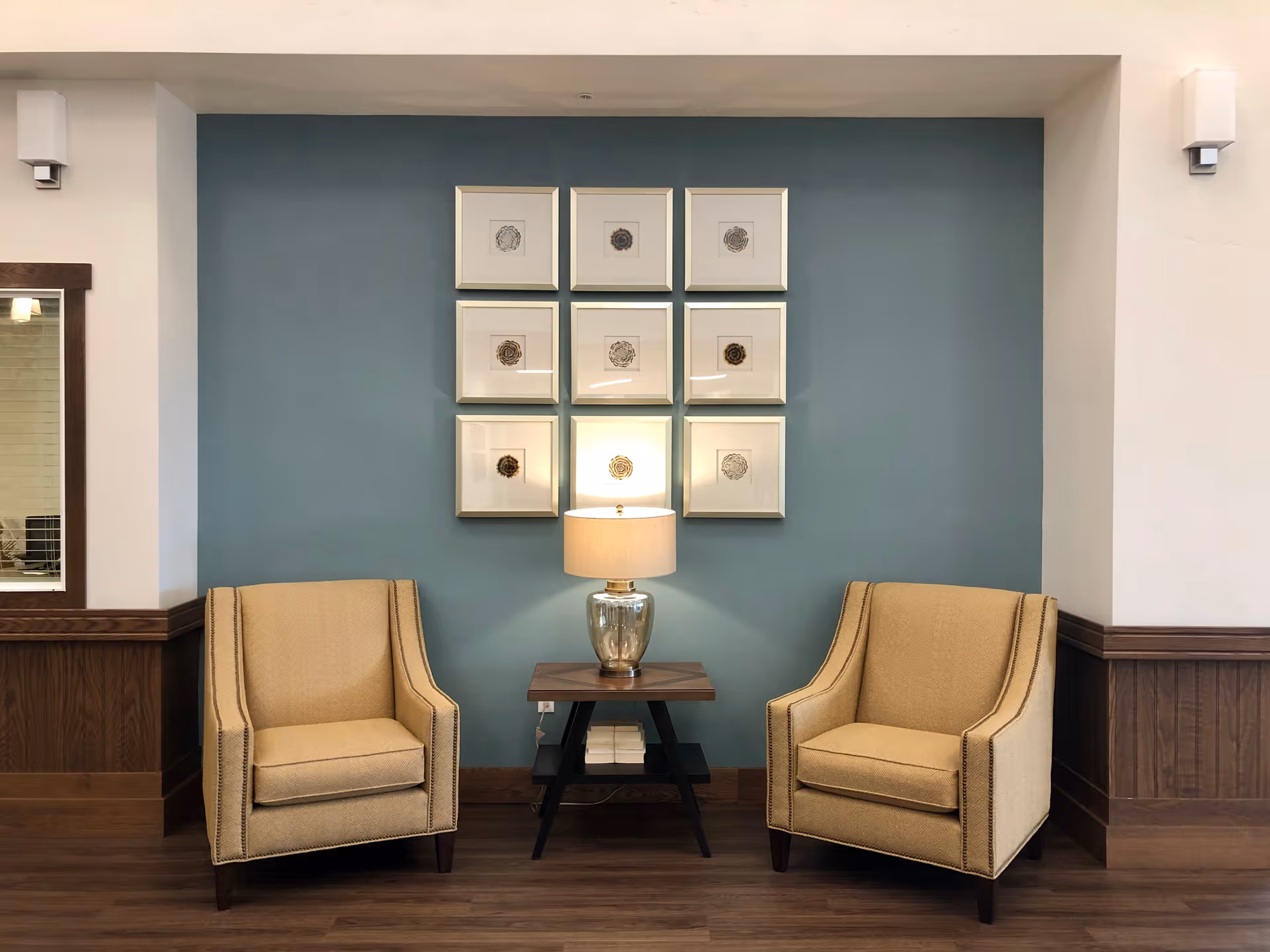 Two beige armchairs flank a small wooden table with a lamp against a teal accent wall hung with nine framed artworks.