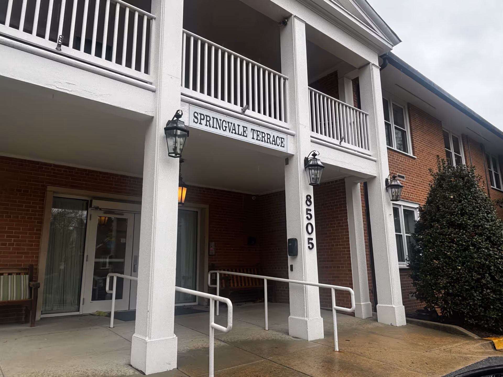 Entrance of a brick building with white pillars and railings, a sign reading 'Springvale Terrace' above the entrance, the number 8505 on one pillar, two lantern-style lights, and benches on either side of the entrance.