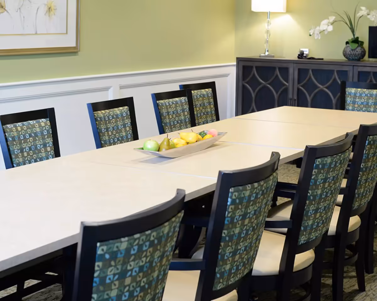 A dining room with a long rectangular table surrounded by chairs with patterned backs and light-colored seats. A decorative bowl with artificial fruit is placed in the center of the table. In the background, there is a dark wooden cabinet with a lamp and a vase with white flowers on top. The walls are painted light green with white wainscoting.