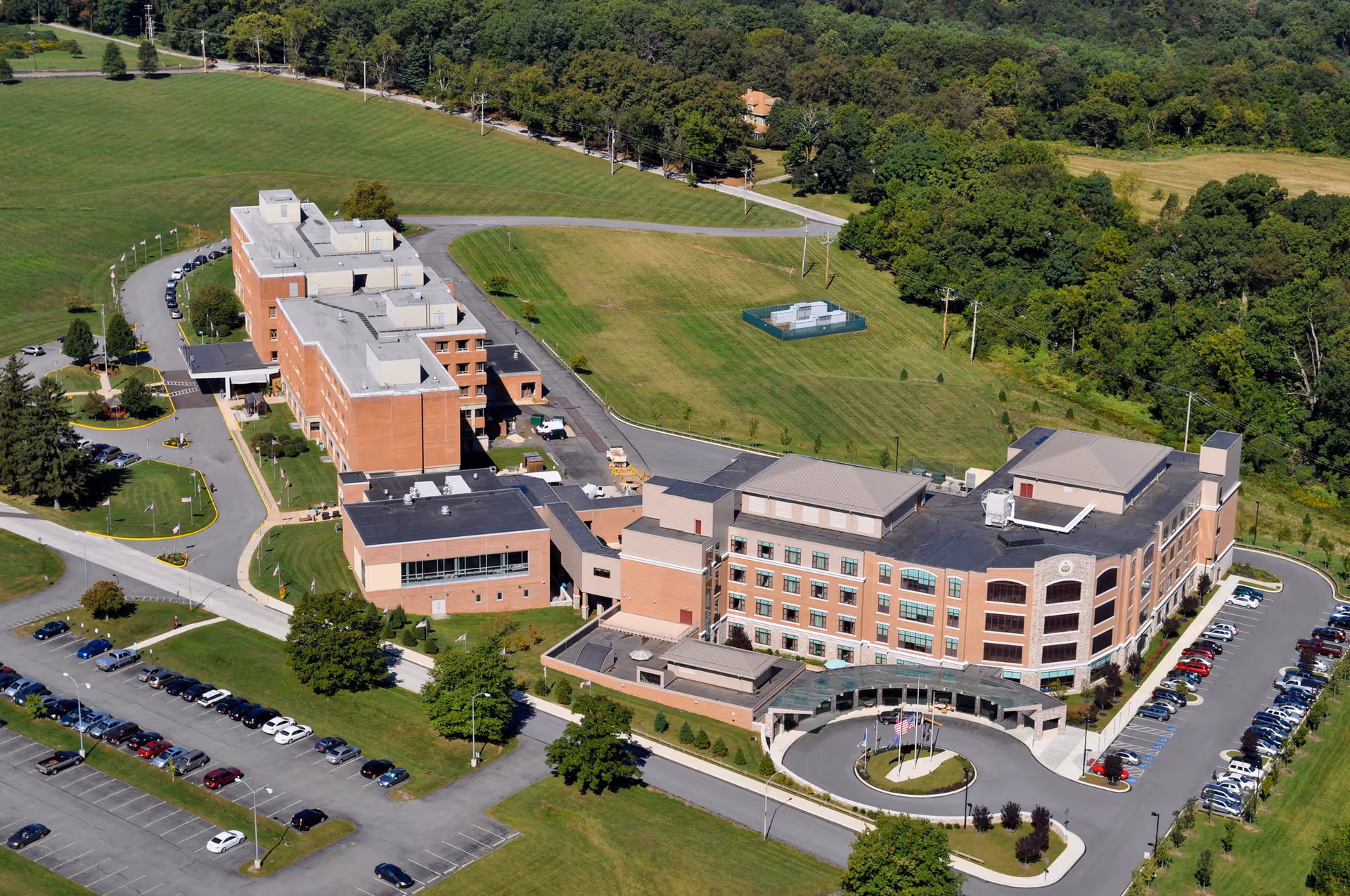 Aerial view of Southeastern Veterans Center showing multiple connected brick buildings surrounded by green lawns, parking lots with cars, and trees in the background.