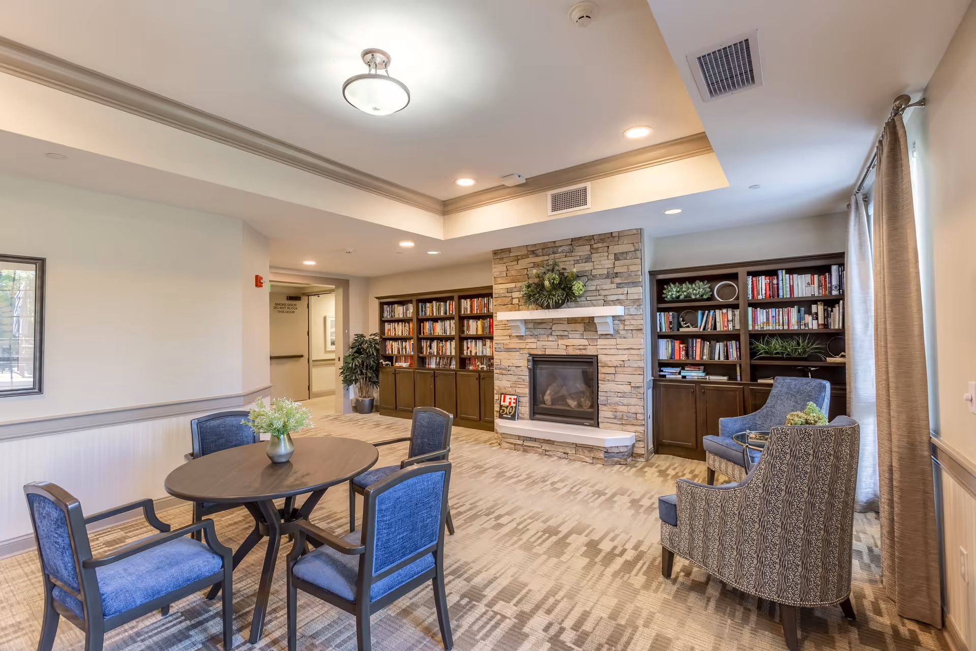A cozy senior living facility common area with a round table and four blue upholstered chairs, a stone fireplace with a mantel decorated with greenery, and built-in bookshelves filled with books and plants. There are two patterned armchairs near a window with beige curtains, and the room is well-lit with ceiling lights.