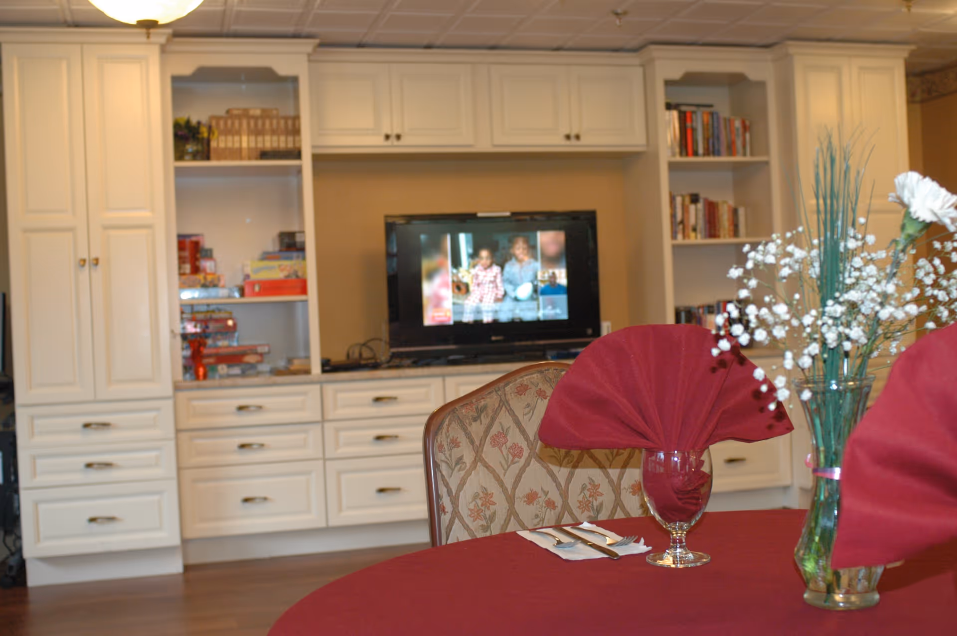 A cozy interior room with white built-in cabinets and shelves, a television displaying a blurred image, and a round table covered with a red tablecloth. The table has two glasses with red folded napkins and a vase with white flowers and green stems.