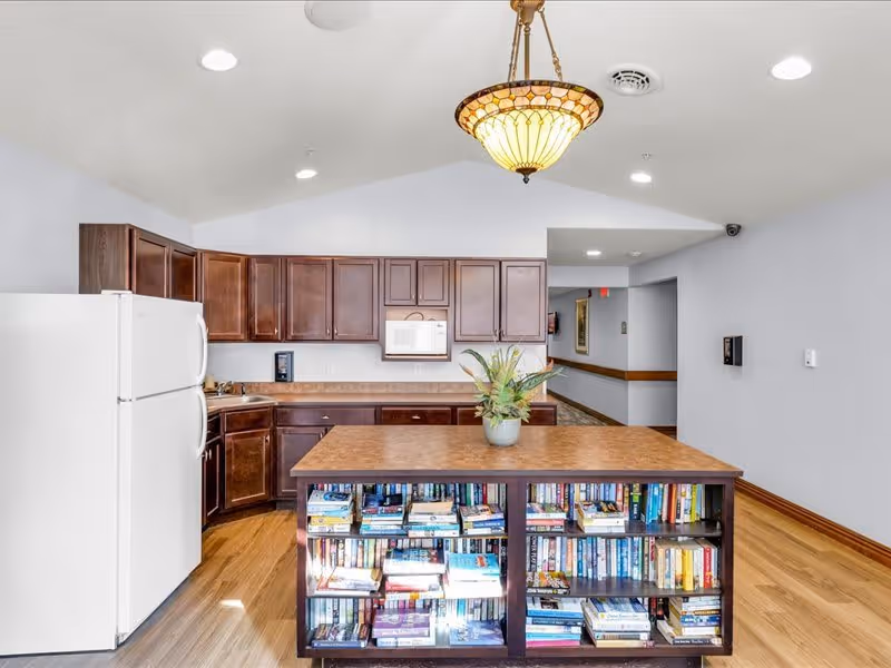 A kitchen area with dark wood cabinets, a white refrigerator, a microwave, and a countertop. In the foreground, there is a kitchen island with a wooden top and shelves filled with books. A decorative hanging light fixture is above the island. The floor is wooden, and the walls are painted light gray.