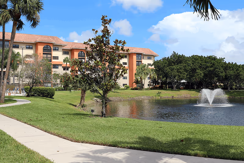 Exterior view of a multi-story senior living building beside a pond with a fountain, walking path, and palm trees.